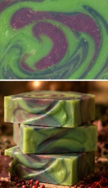 Three stacked rectangular bars of handmade soap with a green and purple marbled swirl pattern, shown both as a close-up top view and stacked on a rustic surface with dried flowers and berries.