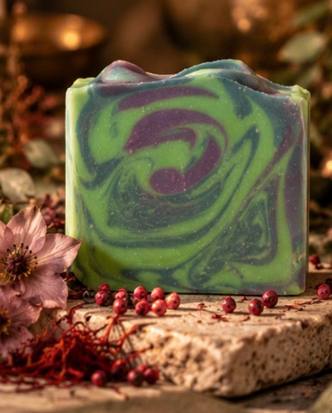 A rectangular handmade soap bar with a green and purple marbled swirl pattern, styled on a stone slab with dried flowers, red berries, and a warm, blurred background.