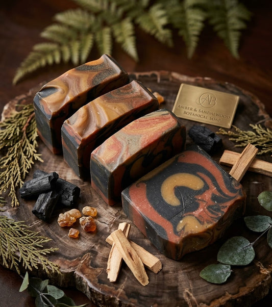Four artisan soap bars with a marbled swirl pattern of black, rust red, orange, and brown, arranged on a rustic wood slice with botanical and wood props. A brass plaque on the wood reads 'AV & SANDWICH BOTANICAL SANCTUARY'.