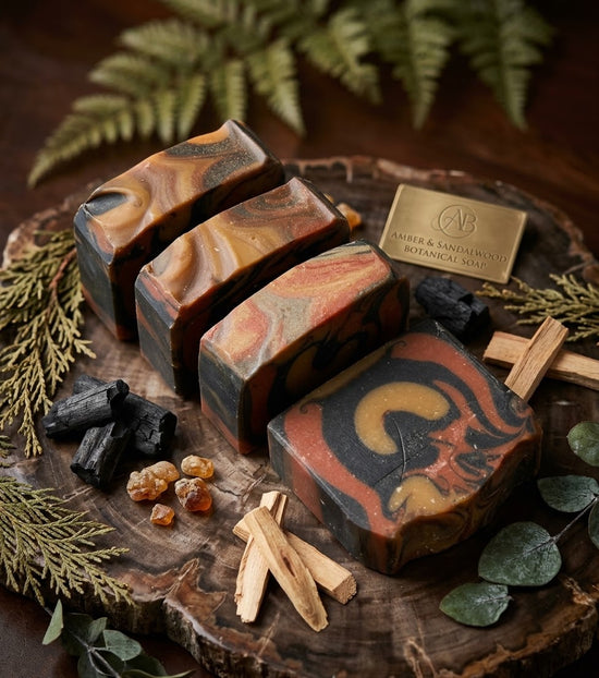 Four artisan soap bars with a marbled swirl pattern of black, rust red, orange, and brown, arranged on a rustic wood slice with botanical and wood props. A brass plaque on the wood reads 'AV & SANDWICH BOTANICAL SANCTUARY'.