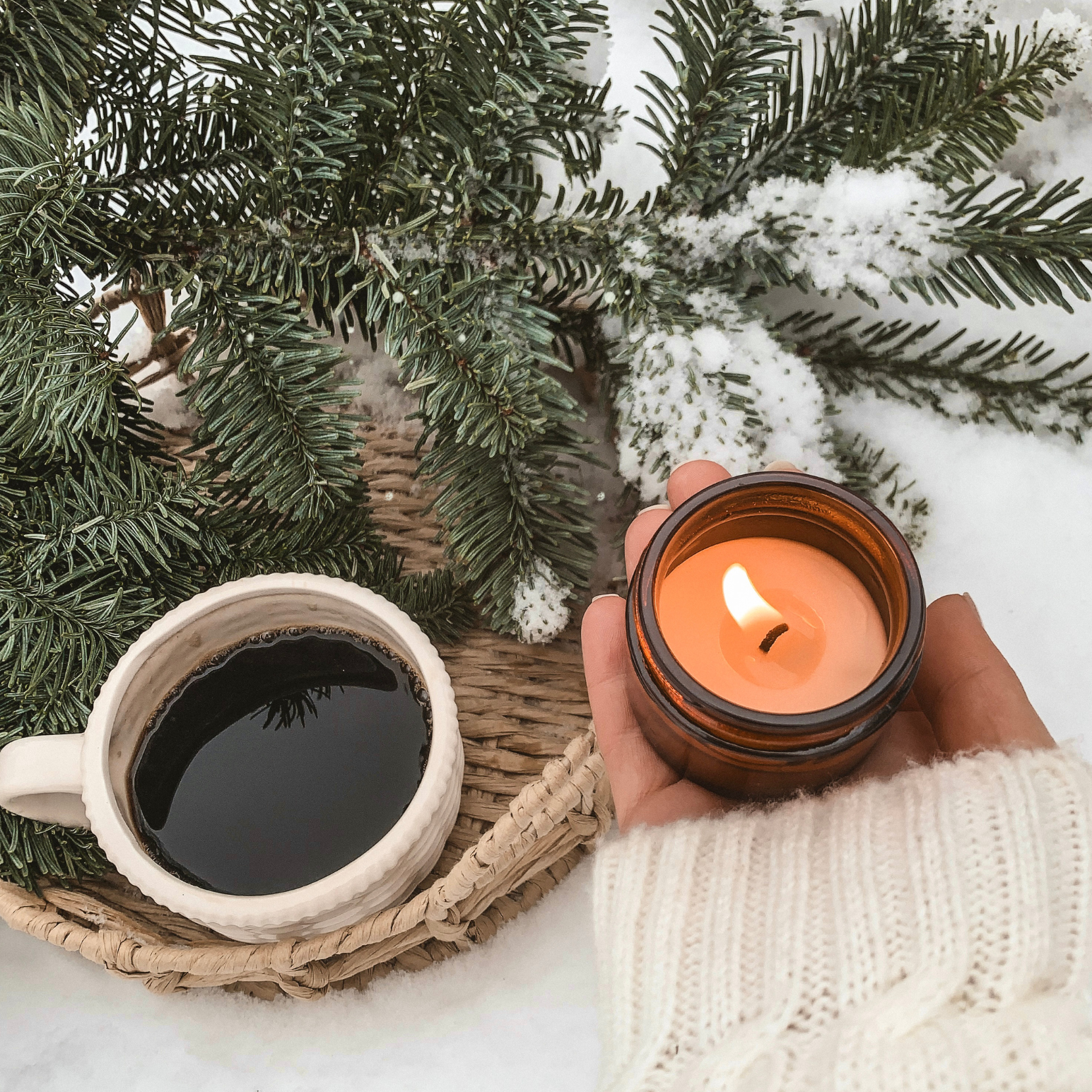 Hand holding a lit candle next to a cup of coffee under snow-covered branches